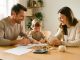 Un homme, une femme et leur jeune enfant organisent un budget familial autour d’une table lumineuse avec des documents vierges, des pièces et une tirelire.